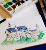 A detailed watercolor painting of a castle with blue roofs, surrounded by lush green foliage at the base. The painting is situated on a piece of paper next to a watercolor palette filled with various colors and brushes, suggesting a creative workspace.