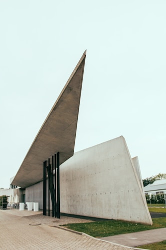 An architectural structure with a sharp, angular roof supported by slender black beams. The building features smooth, grey concrete walls extending horizontally, creating a minimalist and modern appearance. The surrounding environment includes a paved area and patches of greenery.