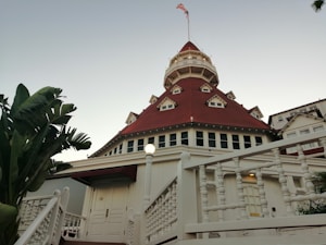 A grand, historic hotel building with a distinctive red roof and multiple gables, set against a clear sky. The structure features detailed woodwork, a prominent tower with windows, and an American flag at its peak. Lush green palm trees frame the scene on the left side.