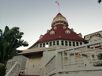 A grand, historic hotel building with a distinctive red roof and multiple gables, set against a clear sky. The structure features detailed woodwork, a prominent tower with windows, and an American flag at its peak. Lush green palm trees frame the scene on the left side.