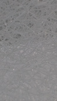 A complex network of tangled, translucent threads or fibers intertwined in an intricate pattern. The strands create an abstract and organized chaos with a smooth and reflective surface.