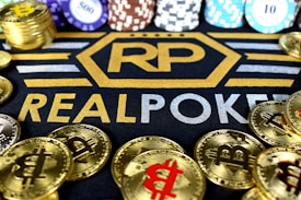 Stacks of poker chips in various colors are arranged in the background, while in the foreground, there are numerous golden coins featuring the Bitcoin symbol. The text 'REALPOKER' is prominently displayed in gold and silver lettering on a dark background.