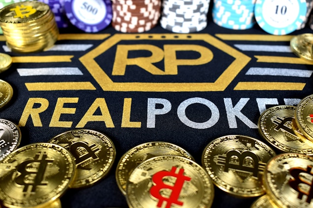 Stacks of poker chips in various colors are arranged in the background, while in the foreground, there are numerous golden coins featuring the Bitcoin symbol. The text 'REALPOKER' is prominently displayed in gold and silver lettering on a dark background.