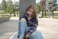 A young person sits on a concrete platform outdoors, leaning against a stone pillar. They are wearing a plaid shirt and ripped jeans, with one knee up. Their expression is thoughtful, with their hand resting against their face. The setting is a park-like environment with trees and pathways, and there is a bright, natural light.