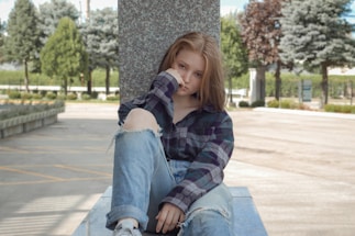 A young person sits on a concrete platform outdoors, leaning against a stone pillar. They are wearing a plaid shirt and ripped jeans, with one knee up. Their expression is thoughtful, with their hand resting against their face. The setting is a park-like environment with trees and pathways, and there is a bright, natural light.