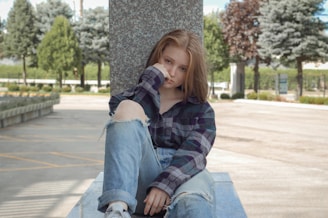 A young person sits on a concrete platform outdoors, leaning against a stone pillar. They are wearing a plaid shirt and ripped jeans, with one knee up. Their expression is thoughtful, with their hand resting against their face. The setting is a park-like environment with trees and pathways, and there is a bright, natural light.