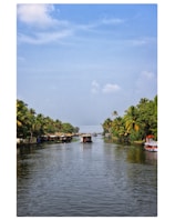 A serene waterway lined with lush green palm trees on both sides. Several small houses and huts are visible near the water's edge. A few boats, including a houseboat, are floating on the calm water. The sky is partly cloudy, adding to the tranquil ambiance.