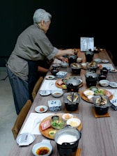 An elderly person is setting a long wooden table with a variety of traditional Japanese dishes, including grilled fish, small bowls of soup, soy sauce, and vegetables. The tableware includes small plates and bowls with different designs, and each setting seems meticulously arranged. The setting suggests a formal or special meal, possibly in a restaurant or home setting.