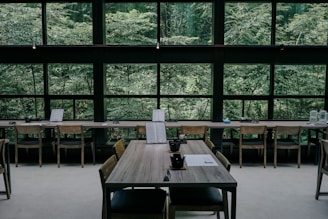 A serene interior of a cafe or restaurant featuring wooden tables and chairs, positioned against large windows that reveal a lush, green forest outside. The tables are set with black teacups and paper menus, creating a minimalistic and calm atmosphere.