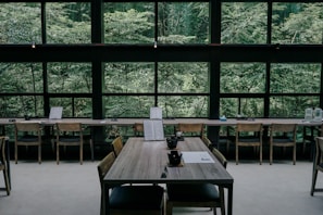 A serene interior of a cafe or restaurant featuring wooden tables and chairs, positioned against large windows that reveal a lush, green forest outside. The tables are set with black teacups and paper menus, creating a minimalistic and calm atmosphere.