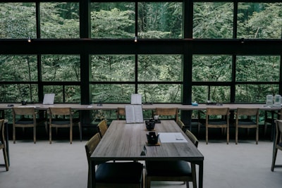 A serene interior of a cafe or restaurant featuring wooden tables and chairs, positioned against large windows that reveal a lush, green forest outside. The tables are set with black teacups and paper menus, creating a minimalistic and calm atmosphere.