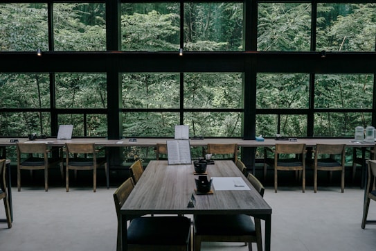 A serene interior of a cafe or restaurant featuring wooden tables and chairs, positioned against large windows that reveal a lush, green forest outside. The tables are set with black teacups and paper menus, creating a minimalistic and calm atmosphere.