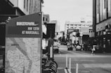 A grayscale urban scene featuring a Biketown map stand in the foreground, indicating its location at NW 13th and Marshall. Behind the stand, a city street stretches into the distance, lined with buildings on both sides. Cars are visible on the street, and a few pedestrians can be seen walking along the sidewalks.