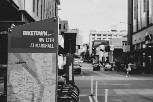 A grayscale urban scene featuring a Biketown map stand in the foreground, indicating its location at NW 13th and Marshall. Behind the stand, a city street stretches into the distance, lined with buildings on both sides. Cars are visible on the street, and a few pedestrians can be seen walking along the sidewalks.