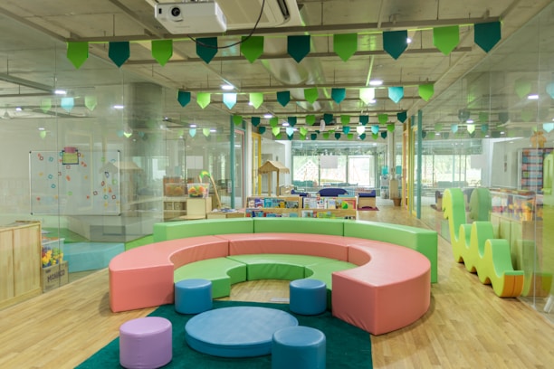 A bright and colorful children's play area with circular seating in pastel shades of pink, green, and blue. The room is filled with various play equipment and toys, including a playhouse and books. The ceiling is decorated with green and blue hanging flags, and the flooring is wooden. Large windows at the far end allow natural light to flood the room.