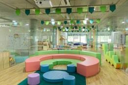 A bright and colorful children's play area with circular seating in pastel shades of pink, green, and blue. The room is filled with various play equipment and toys, including a playhouse and books. The ceiling is decorated with green and blue hanging flags, and the flooring is wooden. Large windows at the far end allow natural light to flood the room.