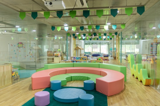 A bright and colorful children's play area with circular seating in pastel shades of pink, green, and blue. The room is filled with various play equipment and toys, including a playhouse and books. The ceiling is decorated with green and blue hanging flags, and the flooring is wooden. Large windows at the far end allow natural light to flood the room.