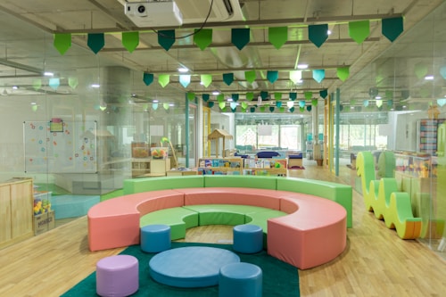 A bright and colorful children's play area with circular seating in pastel shades of pink, green, and blue. The room is filled with various play equipment and toys, including a playhouse and books. The ceiling is decorated with green and blue hanging flags, and the flooring is wooden. Large windows at the far end allow natural light to flood the room.