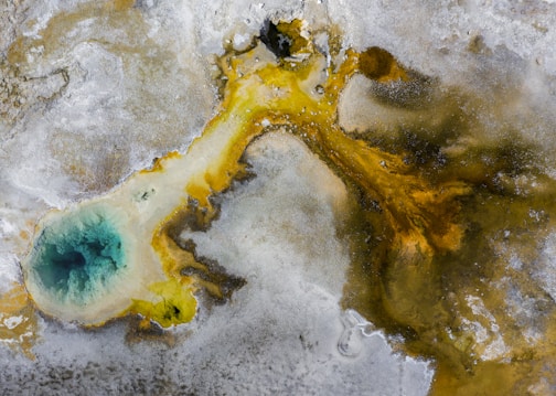 An aerial view of a geothermal area with a vibrant blue pool surrounded by mineral deposits. The striking contrast between the deep turquoise water and the earthy hues of yellow, brown, and gray creates a visually captivating scene. The pool appears to be a hot spring or fumarole, typical of geothermal landscapes.
