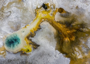 An aerial view of a geothermal area with a vibrant blue pool surrounded by mineral deposits. The striking contrast between the deep turquoise water and the earthy hues of yellow, brown, and gray creates a visually captivating scene. The pool appears to be a hot spring or fumarole, typical of geothermal landscapes.