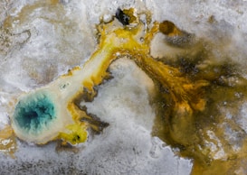 An aerial view of a geothermal area with a vibrant blue pool surrounded by mineral deposits. The striking contrast between the deep turquoise water and the earthy hues of yellow, brown, and gray creates a visually captivating scene. The pool appears to be a hot spring or fumarole, typical of geothermal landscapes.