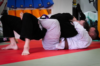 Two people are practicing Brazilian Jiu-Jitsu on a red mat, with one person applying a submission hold on the other. They both wear traditional gi uniforms; one wears a white gi, and the other a black gi. In the background, training pads in blue and orange colors are visible, along with gym equipment.