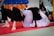 Two people are practicing Brazilian Jiu-Jitsu on a red mat, with one person applying a submission hold on the other. They both wear traditional gi uniforms; one wears a white gi, and the other a black gi. In the background, training pads in blue and orange colors are visible, along with gym equipment.