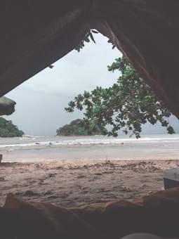 A view from inside a tent looking out towards a beach. The foreground shows the sandy ground and part of the tent's opening. In the background, there are waves on the ocean and an island covered with lush greenery. Tree branches hang down from the top frame of the image, adding a touch of nature. The sky is overcast, indicating a cloudy day.