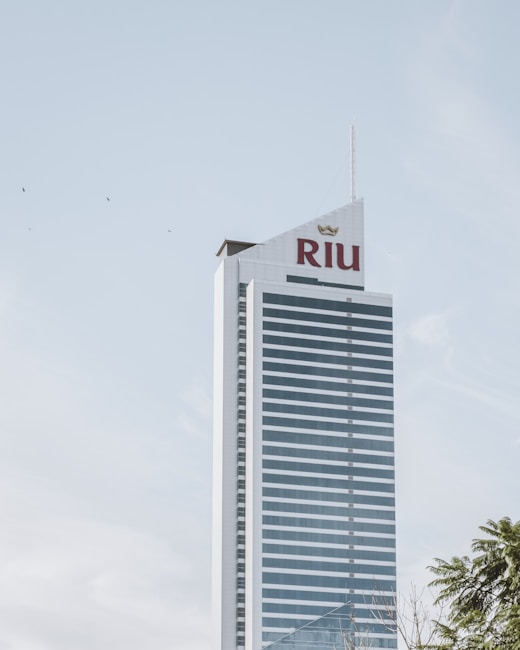 A tall, modern skyscraper with a glass facade and the word 'Riu' displayed prominently in large red letters near the top. The building is set against a clear, pale blue sky with a few birds flying nearby. The bottom right corner reveals the tops of some trees, suggesting an urban setting.