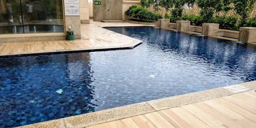 A rectangular swimming pool with clear blue water, surrounded by a beige tiled patio. There is a line of greenery, including bushes and hedges, along one side, providing a natural backdrop. The setting appears to be part of an outdoor area adjacent to a building with glass doors.
