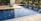 A rectangular swimming pool with clear blue water, surrounded by a beige tiled patio. There is a line of greenery, including bushes and hedges, along one side, providing a natural backdrop. The setting appears to be part of an outdoor area adjacent to a building with glass doors.
