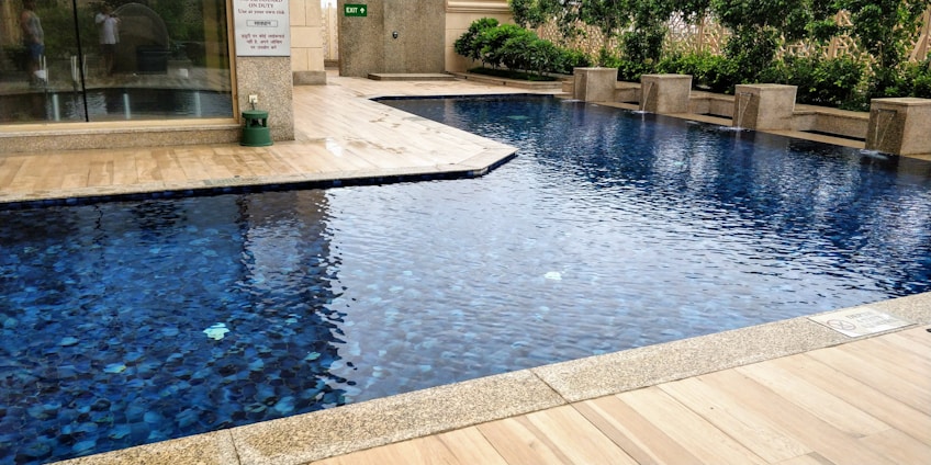 A rectangular swimming pool with clear blue water, surrounded by a beige tiled patio. There is a line of greenery, including bushes and hedges, along one side, providing a natural backdrop. The setting appears to be part of an outdoor area adjacent to a building with glass doors.