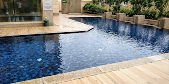 A rectangular swimming pool with clear blue water, surrounded by a beige tiled patio. There is a line of greenery, including bushes and hedges, along one side, providing a natural backdrop. The setting appears to be part of an outdoor area adjacent to a building with glass doors.