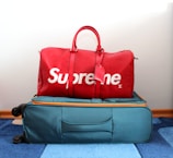 A red duffel bag with the brand name displayed in large white letters sits atop a teal suitcase with orange accents. The suitcase is on a blue and white striped carpet.