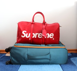 A red duffel bag with the brand name displayed in large white letters sits atop a teal suitcase with orange accents. The suitcase is on a blue and white striped carpet.