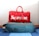 A red duffel bag with the brand name displayed in large white letters sits atop a teal suitcase with orange accents. The suitcase is on a blue and white striped carpet.