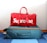 A red duffel bag with the brand name displayed in large white letters sits atop a teal suitcase with orange accents. The suitcase is on a blue and white striped carpet.