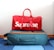 A red duffel bag with the brand name displayed in large white letters sits atop a teal suitcase with orange accents. The suitcase is on a blue and white striped carpet.