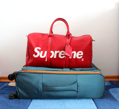 A red duffel bag with the brand name displayed in large white letters sits atop a teal suitcase with orange accents. The suitcase is on a blue and white striped carpet.