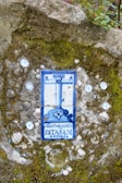 A ceramic tile embedded in a moss-covered stone wall, featuring a blue and white design with what appears to be a crucifix and decorative text. The tile is surrounded by small circular ornaments embedded in the wall.