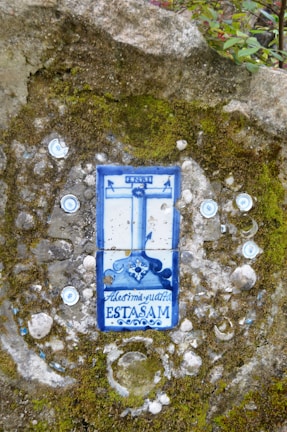 A ceramic tile embedded in a moss-covered stone wall, featuring a blue and white design with what appears to be a crucifix and decorative text. The tile is surrounded by small circular ornaments embedded in the wall.