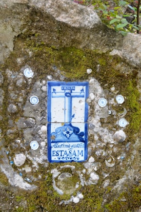 A ceramic tile embedded in a moss-covered stone wall, featuring a blue and white design with what appears to be a crucifix and decorative text. The tile is surrounded by small circular ornaments embedded in the wall.