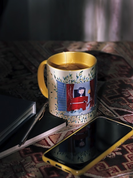 A cup with a colorful design is placed on a patterned fabric surface. Next to it, a pen rests on a closed notebook, while a smartphone with a yellow case lies nearby. The image combines elements of still life with a hint of personal workspace.