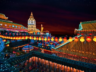 An elaborate temple complex is decorated with numerous colorful lanterns and lights, illuminating the structures against a dark evening sky. The architecture includes ornate roofs and a tall, intricately designed tower, all adorned with glowing ornaments and festive lighting.