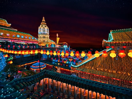 An elaborate temple complex is decorated with numerous colorful lanterns and lights, illuminating the structures against a dark evening sky. The architecture includes ornate roofs and a tall, intricately designed tower, all adorned with glowing ornaments and festive lighting.