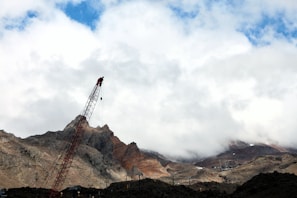 A towering crane stands against a backdrop of rugged mountains partially shrouded in clouds, with hints of blue sky peeking through. The crane is angled and positioned near the base of the mountains, which display a mix of brown, gray, and red hues.