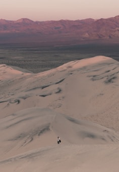 A vast desert landscape features rolling sand dunes in the foreground with a solitary person walking at the base. The background showcases a range of distant mountains under a soft, pinkish sky, creating a sense of depth and isolation.