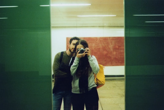 A couple is taking a photo through a glass surface, with one person holding a camera. They are standing close together, and the background includes a large red panel and a green reflective surface.