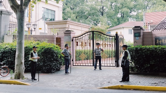 Four security guards stand at the entrance of a large gated property, each positioned at different points around the entrance. The gate is ornately designed with gold elements. There is a tree on the left side, and lush green hedges surround the area. In the background, a building and another residential structure are visible.
