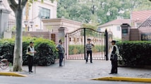 Four security guards stand at the entrance of a large gated property, each positioned at different points around the entrance. The gate is ornately designed with gold elements. There is a tree on the left side, and lush green hedges surround the area. In the background, a building and another residential structure are visible.